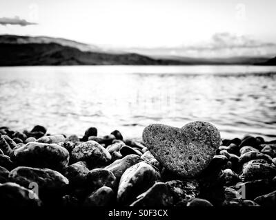 Summer love; in black and white. Heart shaped rock on the beach with lake and mountains in the background. Stock Photo