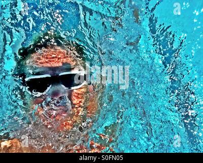 Closeup view above male swimmer doing backstroke facing up in an outdoor swimming pool competition. Sunlit wake, waves and bubbles. Stock Photo