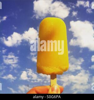 Orange ice-lolly on a background of deep blue sky with fluffy white clouds Stock Photo