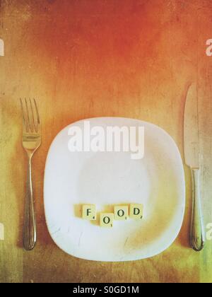 It's a photo of square scrabble letters, placed on a dinner plate, with cutlery on a wooden table, forming the word FOOD. Stock Photo