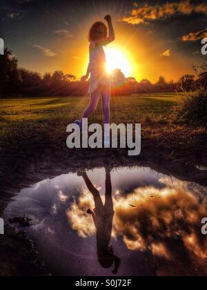 Young girl posing with raised arm in front of setting sun reflected in puddle Stock Photo