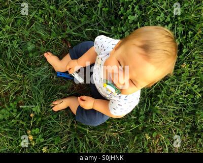 View from the top of a 8 months old smiling  baby boy sitting outside on the ground, green grass with pacifier in his mouth, holding a clothes pin Stock Photo