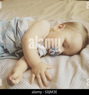 8 month old baby boy sleeping peacefully on a hot summer day on parents bed covered with a light blue blanket, with a pacifier in his mouth Stock Photo