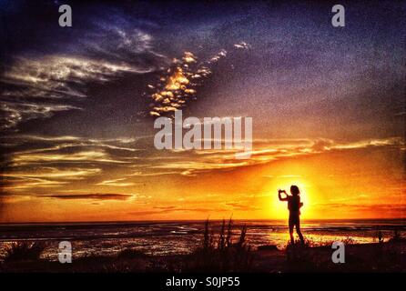 Young girl taking photo silhouetted against the setting sun over Morecambe Bay at Heysham Stock Photo