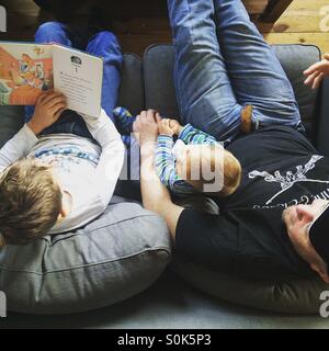 Photo from above of a father with his two children , on of whom is reading a book aloud to his younger brother , sitting on a grey couch. Stock Photo