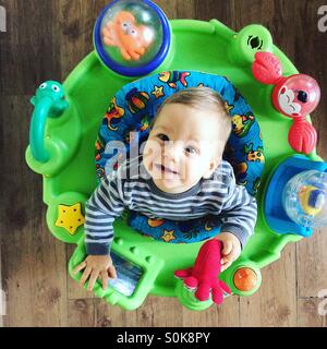 Photo from above  of a smiling 9 months old baby boy , who is sitting in his toy jumpy chair , having a great time, twirling around being  surrounded by different toys