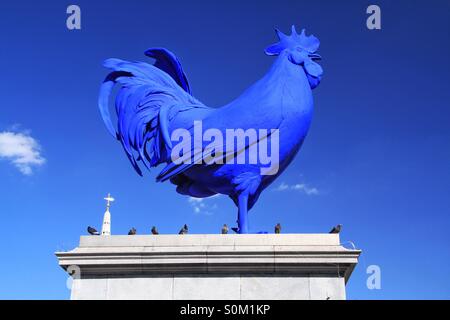 A big, stone, blue chicken statue sits on top of a plinth in Trafalgar Square in London with a blue sky behind it and some pigeons as company. Stock Photo