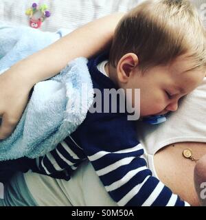 Tired 9 months old baby boy taking a nap on top of his mom . Stock Photo