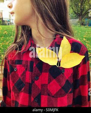 A young girl wearing Red and black buffalo-checked plaid shirt has 3 yellow leaves in her shirt pocket. Stock Photo