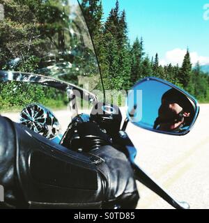 Motorist wearing a black leather jacket riding a Harley Davidson cruising on the streets of Jasper, Canada, lined with pine trees accompanied by blue skies. Stock Photo