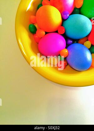 A bowl of brightly dyed eggs and jelly beans in a bowl viewed from above on white background