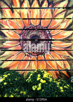 A bright orange and yellow flower painted on a wood pallet with yellow flowers in front of it Stock Photo