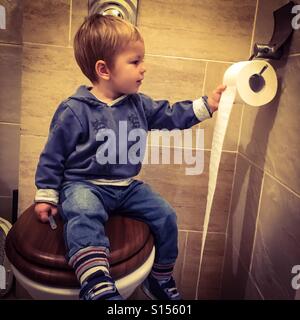 Toddler sitting on a toilet seat , having fun unrolling toilet paper Stock Photo