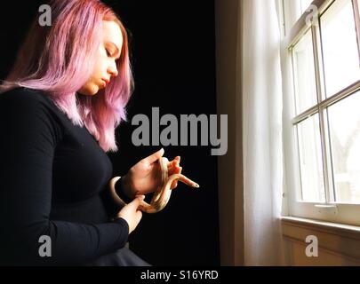 A teenage girl holding a pet snake next to a window. Stock Photo