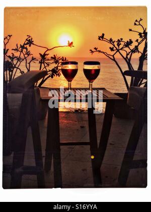 Two Mexican blue rimmed wine glasses are filled with red wine on a teak table between two chairs on a deck overlooking a sunset over the Pacific Ocean in Nayarit, Mexico. Stock Photo