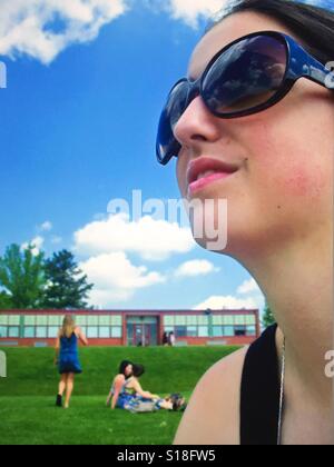A close up profile of a dark haired girl wearing dark sunglasses against a blue sky with clouds and a school and schoolyard in the background Stock Photo