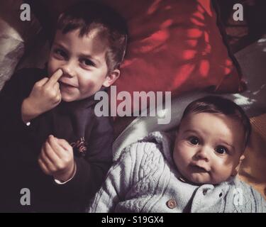 Two brothers lying down next to each other on pillows, one of the boys is picking his nose and the other is just looking straight to camera. Stock Photo