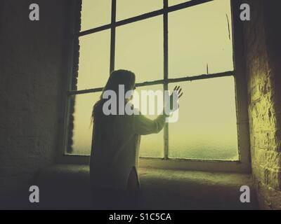 Rear view silhouette of a young woman looking out of the window hand touching the glass Stock Photo