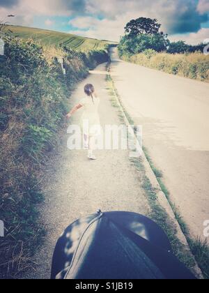 Cute little girl walking to school along a narrow rural Cornish road on a lovely warm sunny morning. Personal perspective of female child from behind. Stock Photo