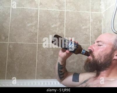 Bald, bearded, tattooed man in hot bath drinking beer from the bottle. Stock Photo