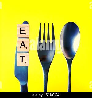 A close-up overhead shot of a knife and a fork and a spoon above a yellow background with the word eat on the knife