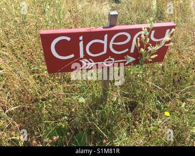 'Cidery' painted in white on handmade red wooden sign with arrow indicating to right. Cider brewery sign in field of long grass Stock Photo