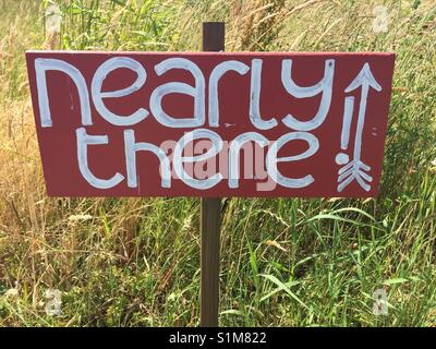 'Nearly there' painted in white on red handmade wooden sign, with exclamation mark and directional arrow. Sign located outdoors with long grass as background Stock Photo