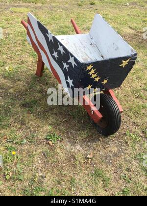 Wooden wheelbarrow with Alaskan and American flags painted on sides in outdoor settling. Grass background Stock Photo