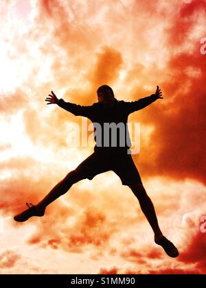 The silhouette of a man jumping high in the air of falling from the sky with a stormy sky behind that looks like an explosion or ball of flames Stock Photo