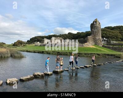 The 12th Century Ogmore Castle, South Wales, is the backdrop to people walking on stepping stones across the river Ogmore Stock Photo