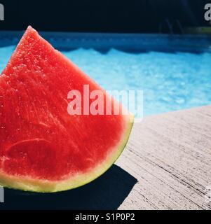 A wedge of juicy red watermelon sitting at the edge of a swimming pool on a sunny day. Stock Photo