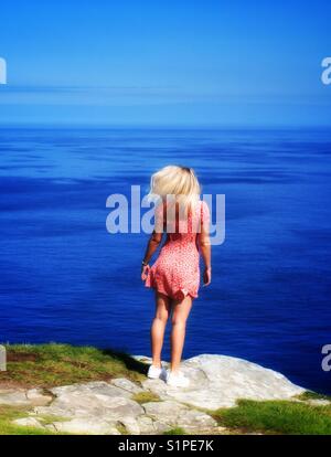 A pretty young blonde haired girl in red floral dress and skirt posing and standing on the edge of a cliff with a deep blue ocean behind on a beautiful summer day Stock Photo