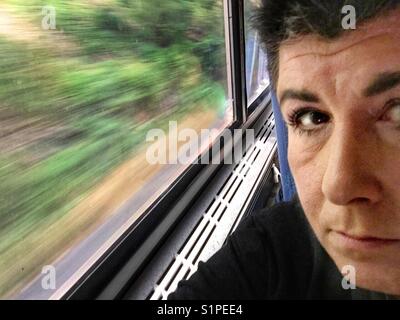 A portrait of a middle aged woman with dark hair who is riding on a train.  The outdoors is seen out the window in motion. Stock Photo