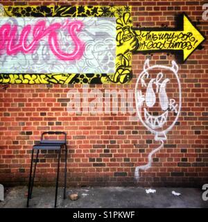 Colorful graffiti on a red brick wall that says “this way to good times” and that has a comical face painted below it.  A tall chair sits against the wAll Stock Photo