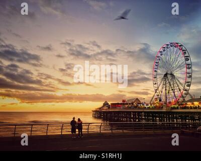 Two people looking out to sea from Blackpool promenade at sunset. Colourful sky and illuminated big wheel on Central Pier Stock Photo