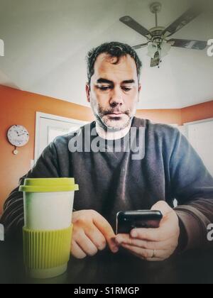 Unshaven 40 year old man reading news articles on his smartphone while drinking a coffee in his kitchen Stock Photo