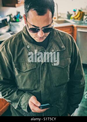 Man standing in kitchen wearing coat and sunglasses using a smartphone Stock Photo