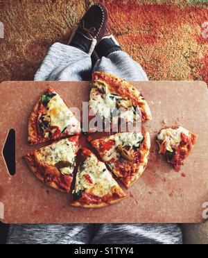 From above view of whole pizza with one partially eaten slice in a cutting board on a person’s lap Stock Photo