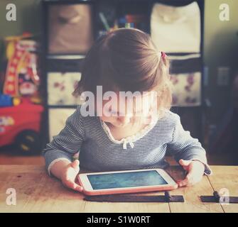 Young toddler girl playing with a tablet in a pink case on a rustic table with family room in background Stock Photo