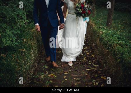 A bride in a beautiful white gown and a groom in a bright blue suit walk down a brick path hand in hand on their wedding day. Stock Photo