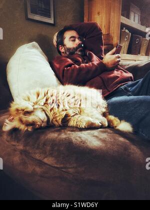 Man with dark beard relaxing on sofa chair using smartphone with large orange cat sleeping in a cute position beside him Stock Photo