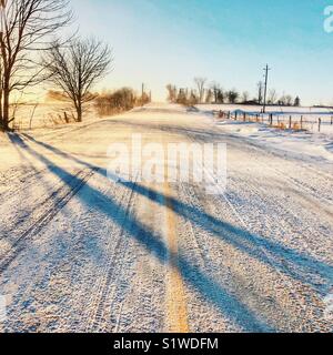 Rural road with yellow line covered in snow with wind blowing snow across at sunset Stock Photo