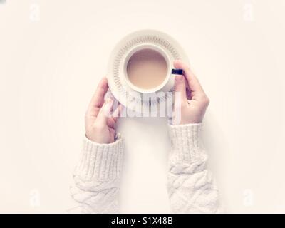 Looking down onto a white background with a girl’s hands holding a cup of tea in a cup and saucer. Stock Photo