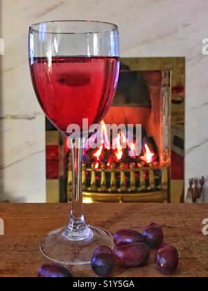 Still life of a glass of red wine and red grapes on a wooden table in front of a coal fire with brass surrounds Stock Photo