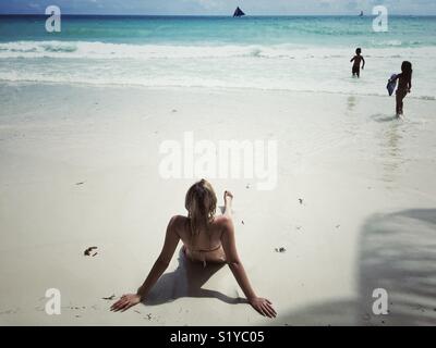 Young woman on white sand beach in bikini watching kids playing in water Stock Photo