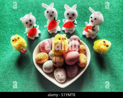 Sugar coated mini chocolate Easter eggs in a heart shaped dish, with mini toy Easter bunnies  and Easter chicks on a green background Stock Photo