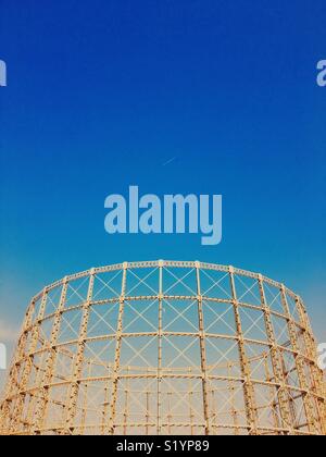Skeleton structure of empty gasometer against a vivid blue sky with jet and contrail Stock Photo