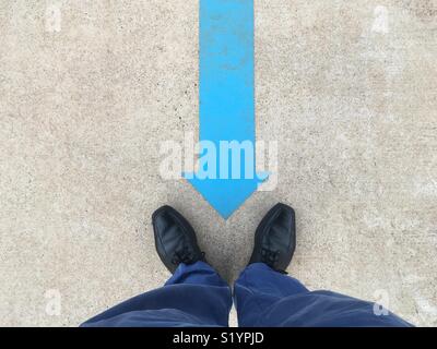 A blue arrow painted on the ground pointing to a man’s black shoes and blue pants. Stock Photo