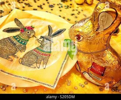 Chocolate Easter bunny wrapped in gold foil paper peering down a paper napkin featuring two Easter rabbits Stock Photo