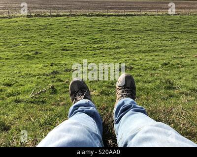 Man hiker sitting on grass with focus on legs wearing blue jeans and hiking shoes with green field meadow in background Stock Photo